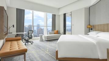 A modern hotel room with a large bed, a sofa, a desk, and a view of the cityscape through large windows.