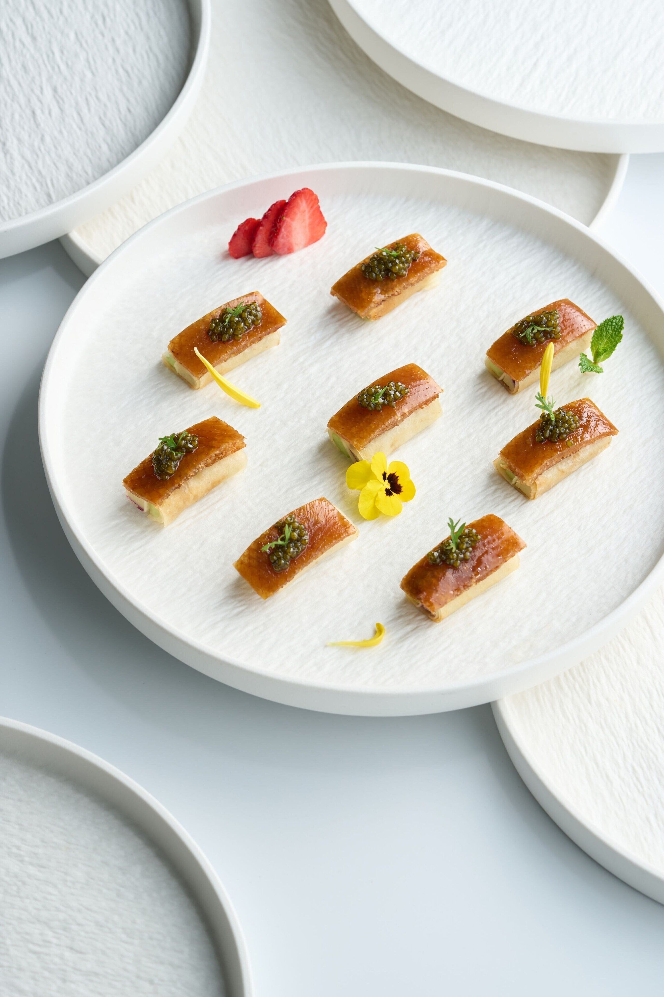 A white plate with small rectangular pastries garnished with herbs and caviar, accompanied by a strawberry and a yellow flower.