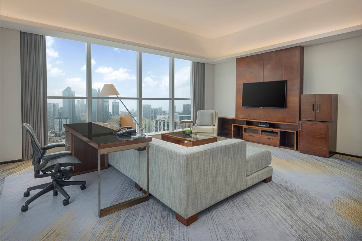 A modern hotel room with a large window offering a city view, a desk with a chair, a sofa, a TV, and a cabinet.