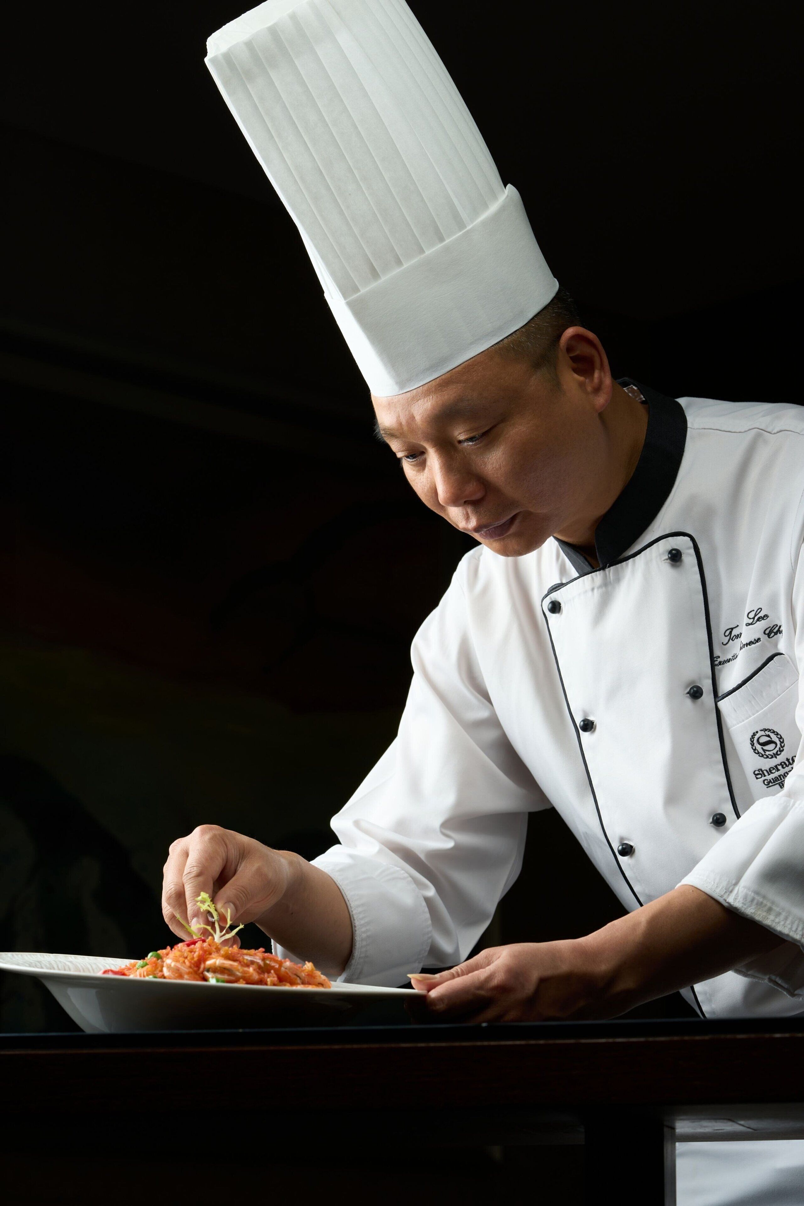 A chef in a white uniform with a tall white hat, preparing food on a plate.