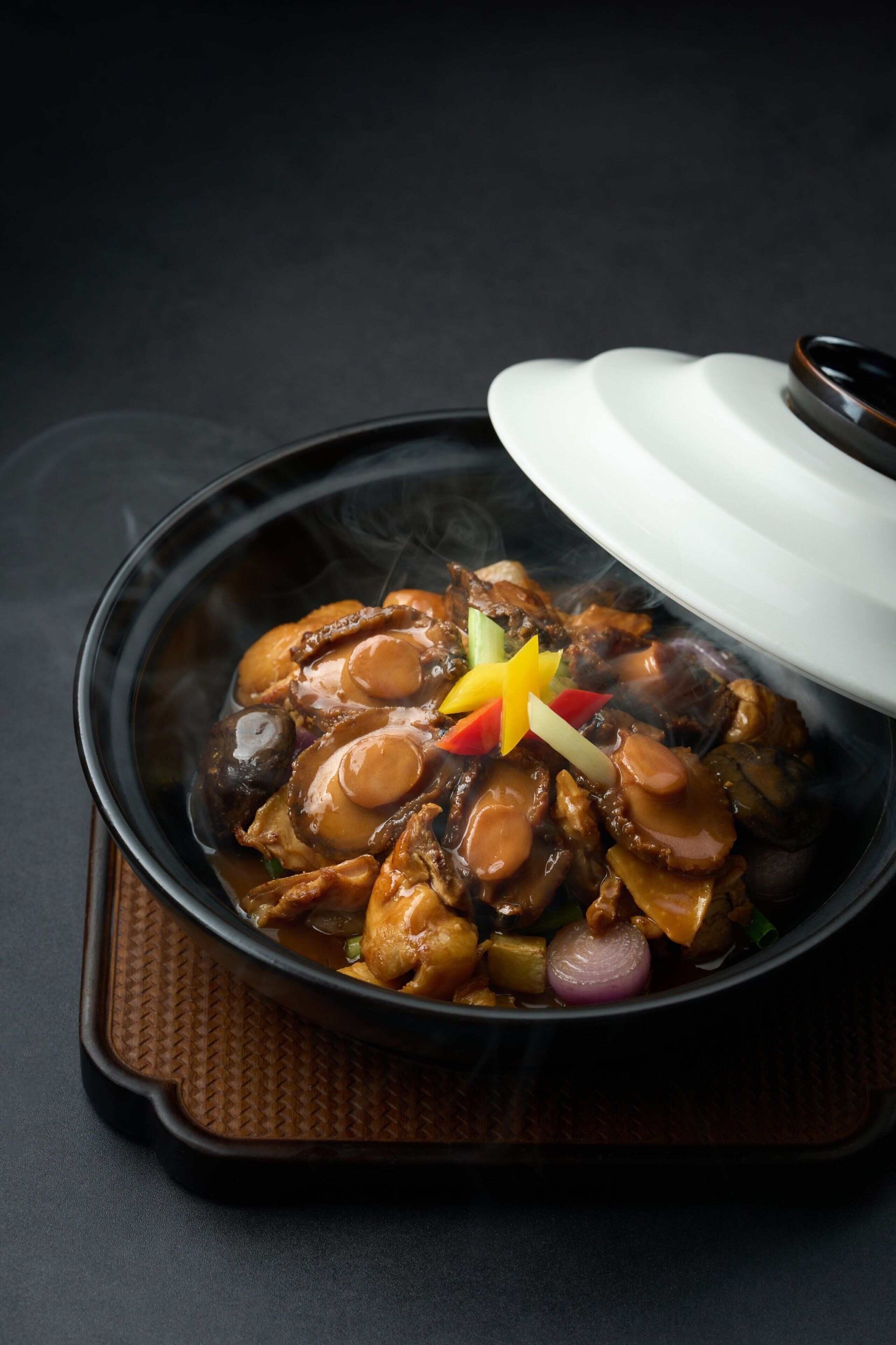 A bowl of food with a lid, steam rising from it, placed on a textured mat.
