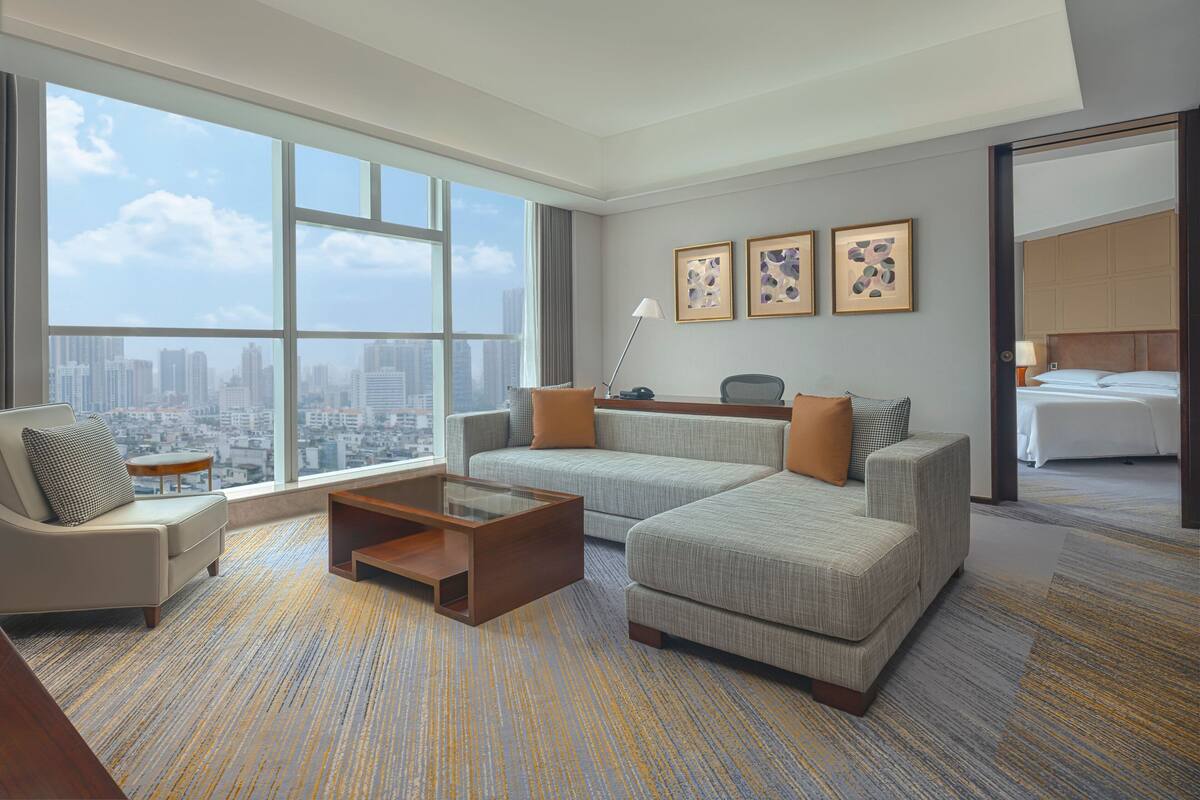 A modern hotel room with a large window offering a city view, a sofa set, a coffee table, and a bed with a headboard.
