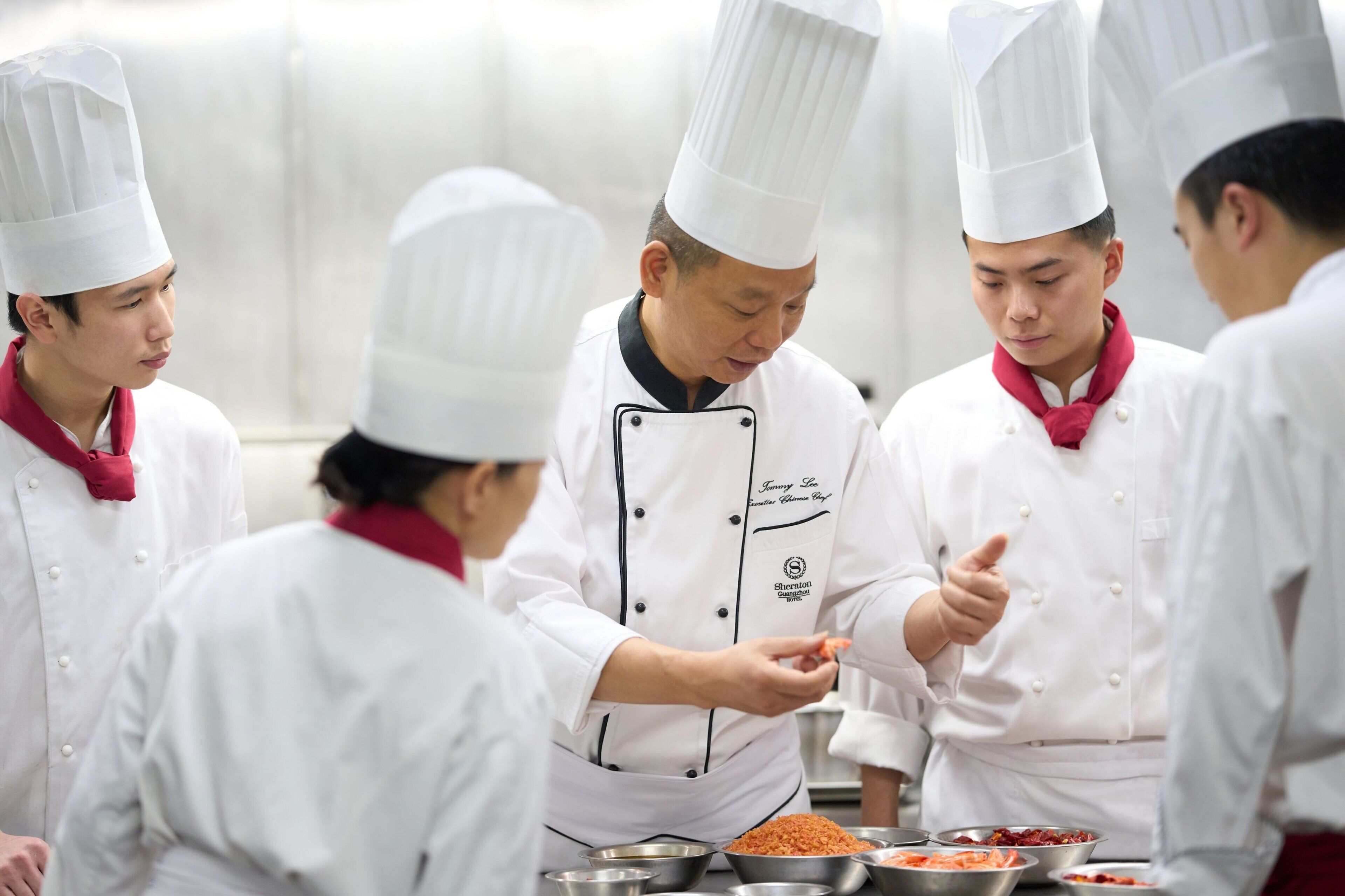 Un groupe de chefs, vêtus d’uniformes blancs rehaussés d’éléments rouges, se tient autour d’une table sur laquelle sont disposés divers aliments.