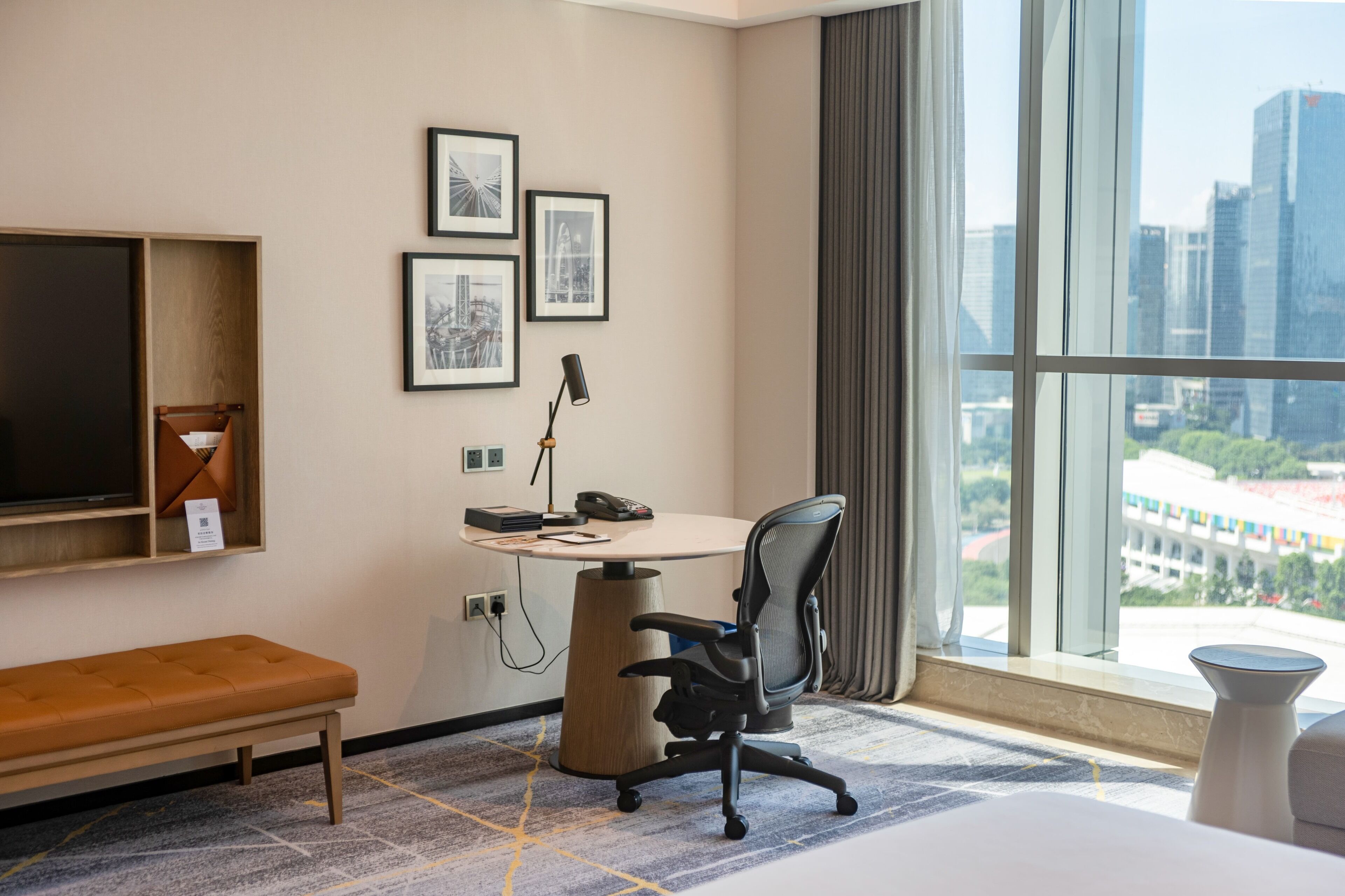 Habitación de hotel moderna con un ventanal grande, un escritorio, una silla, un banco y cuadros en la pared.