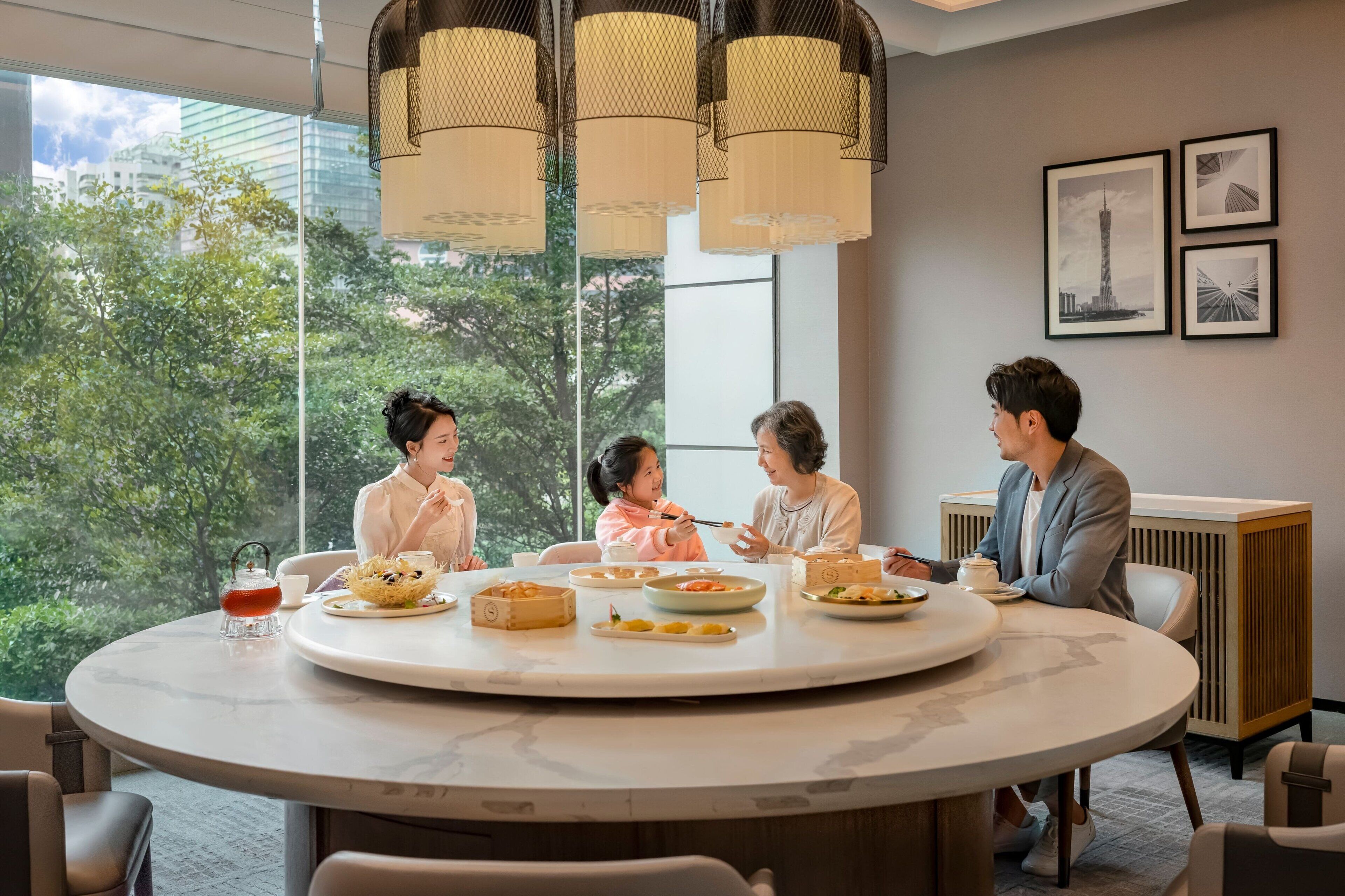A round table with four people seated around it, engaged in conversation. The room has large windows with a view of trees and buildings outside.