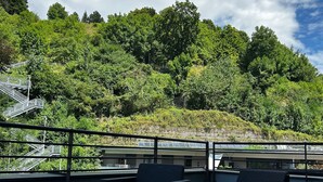 A rooftop terrace with wicker furniture, a metal railing, and a view of a lush green hillside. - Hotel Karwendel (Innsbruck)