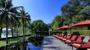 A poolside area with lounge chairs and umbrellas, surrounded by palm trees and a view of a lake. - U Inchantree Kanchanaburi (Kanchanaburi)