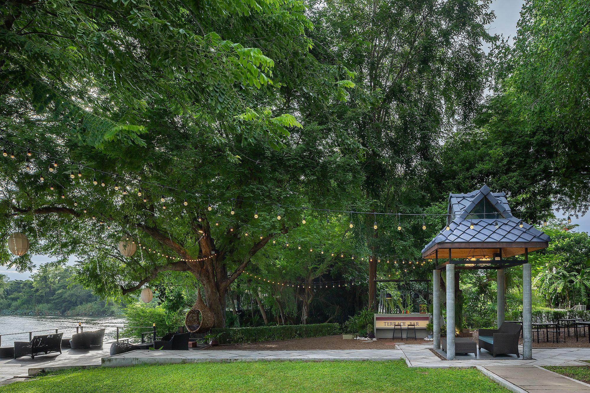 A gazebo with a sign reading 'TERACE BAR' in a park with a large tree, seating, and a view of a body of water.