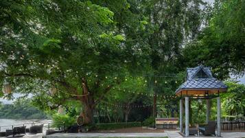 Un kiosque avec une enseigne indiquant « BAR DE PLEIN AIR », situé dans un parc avec un grand arbre, des sièges et une vue sur une étendue d’eau.