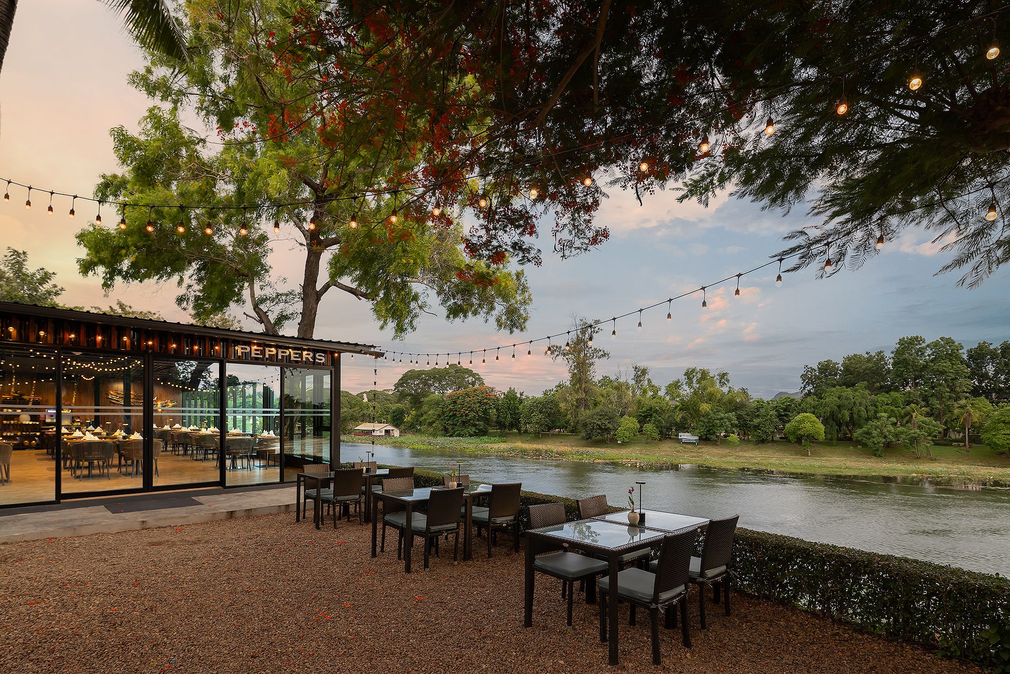 An outdoor dining area with tables and chairs, a glass building with a sign, and a body of water in the background.