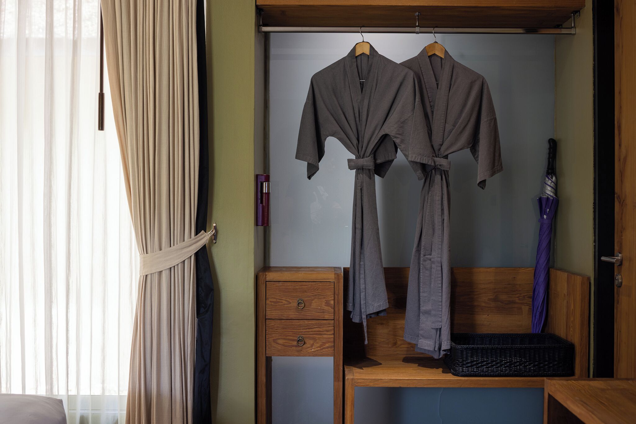 A room with a wooden desk, a small drawer, a wicker basket, and two robes hanging on a rack.