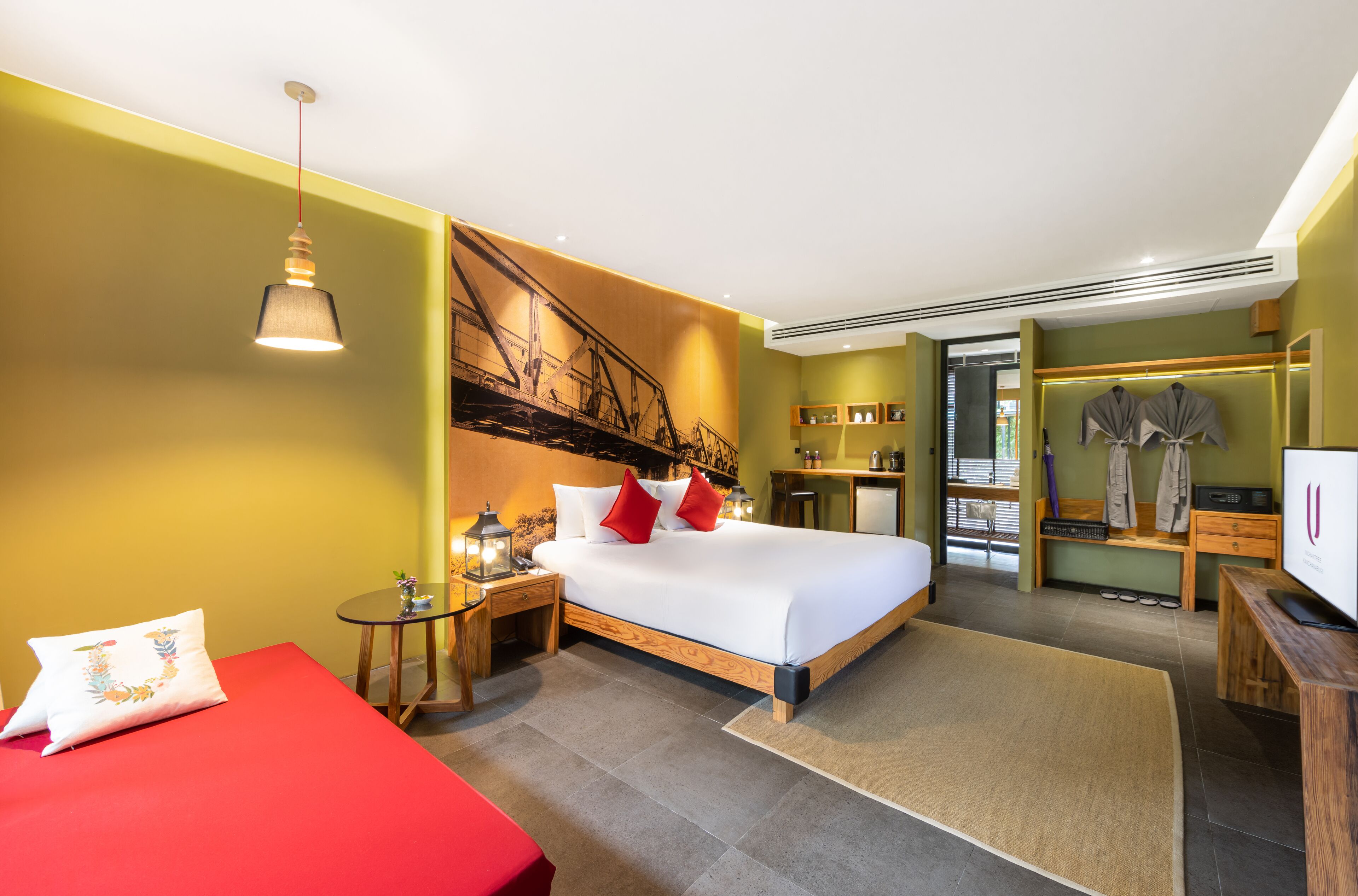 A hotel room with a bed, a red sofa, a small table with a lamp, and a TV.