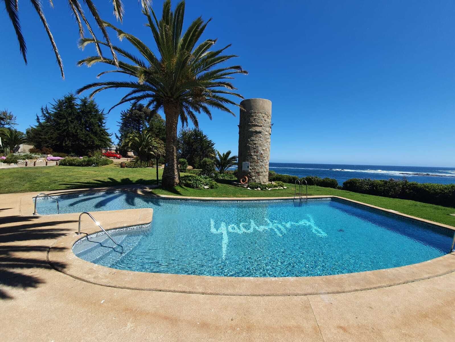 Piscina con cielo azul despejado y vista al océano, rodeada de palmeras y un pilar de piedra.