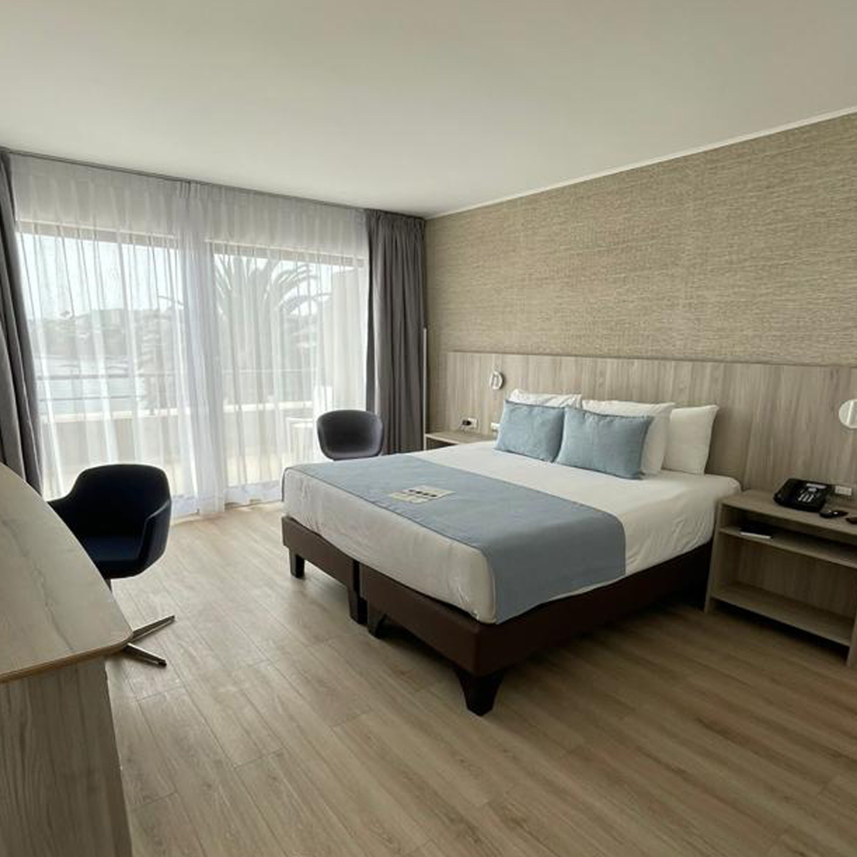 Habitació d'hotel moderna amb un llit gran, una taula de nit amb una làmpada, una cadira i vista a l'exterior a través de cortines translúcides.