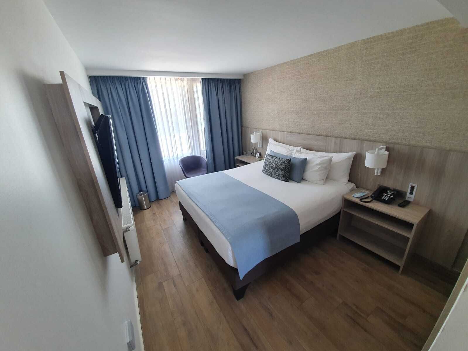 A hotel room with a bed, bedside tables, a desk, a chair, a window with curtains, and a wall-mounted TV.