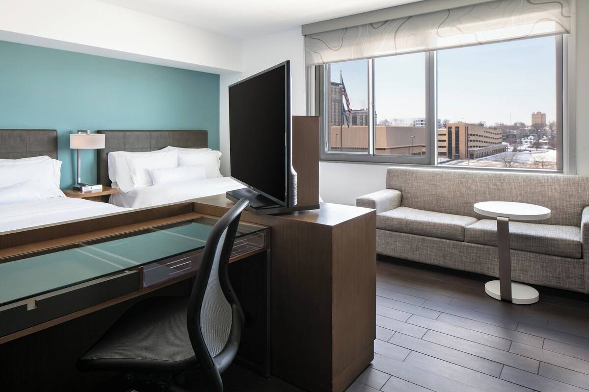 Habitación de hotel con dos camas, un escritorio con computadora, televisión, sofá y vista a la ciudad.
