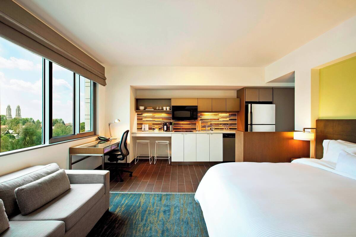 Habitación de hotel con cama, sofá, escritorio con silla, kitchenette y un ventanal con vistas a la ciudad.