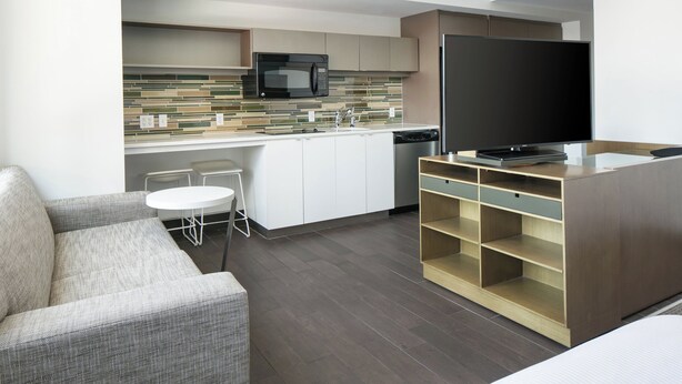 A modern kitchen with a microwave, dishwasher, and a large flat-screen TV. There is a sofa and a small table in the living area. - Element by Marriott Omaha Midtown Crossing (Omaha)
