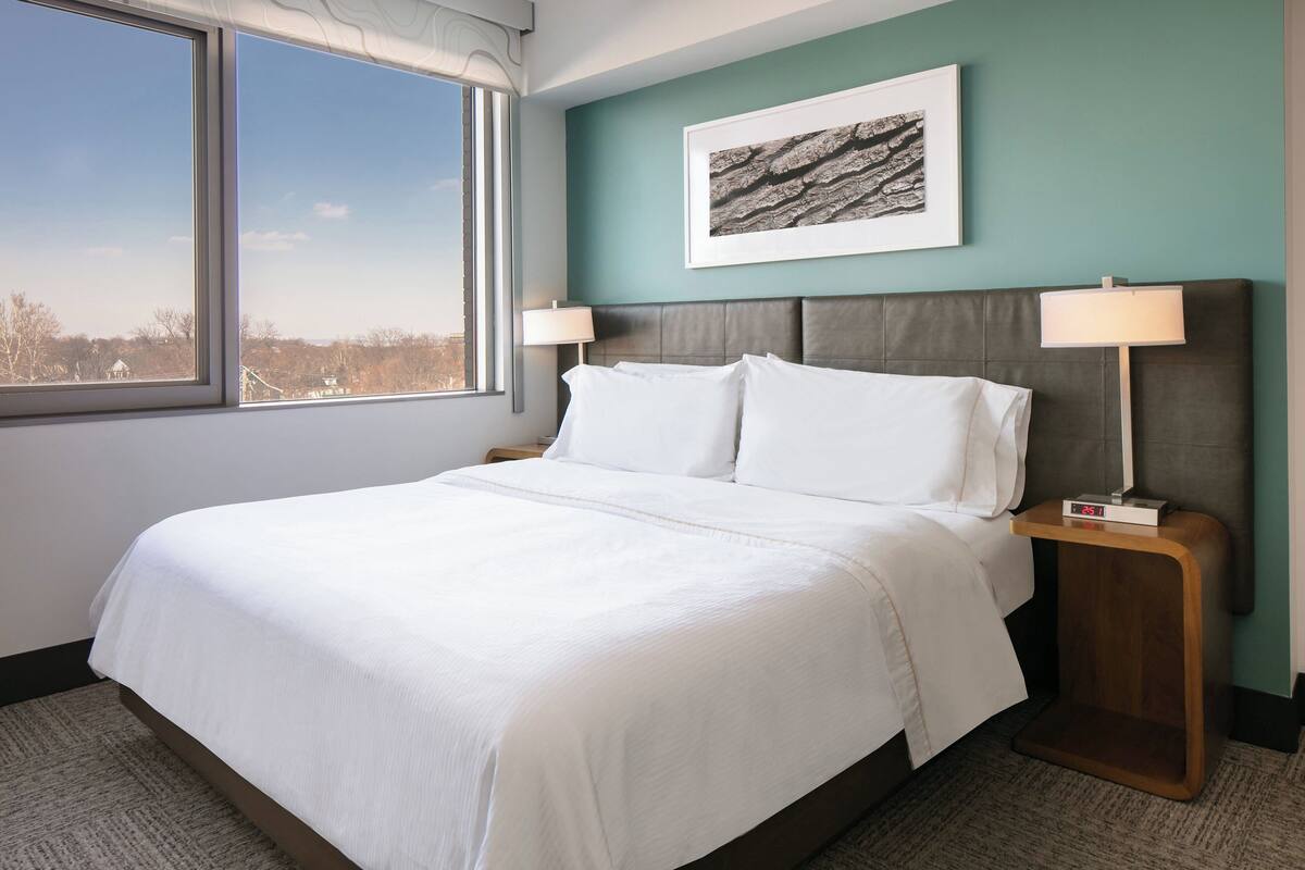 Habitación de hotel con una cama grande, dos mesitas de noche con lámparas, un reloj y un cuadro en la pared, además de vistas al exterior a través de un ventanal amplio.