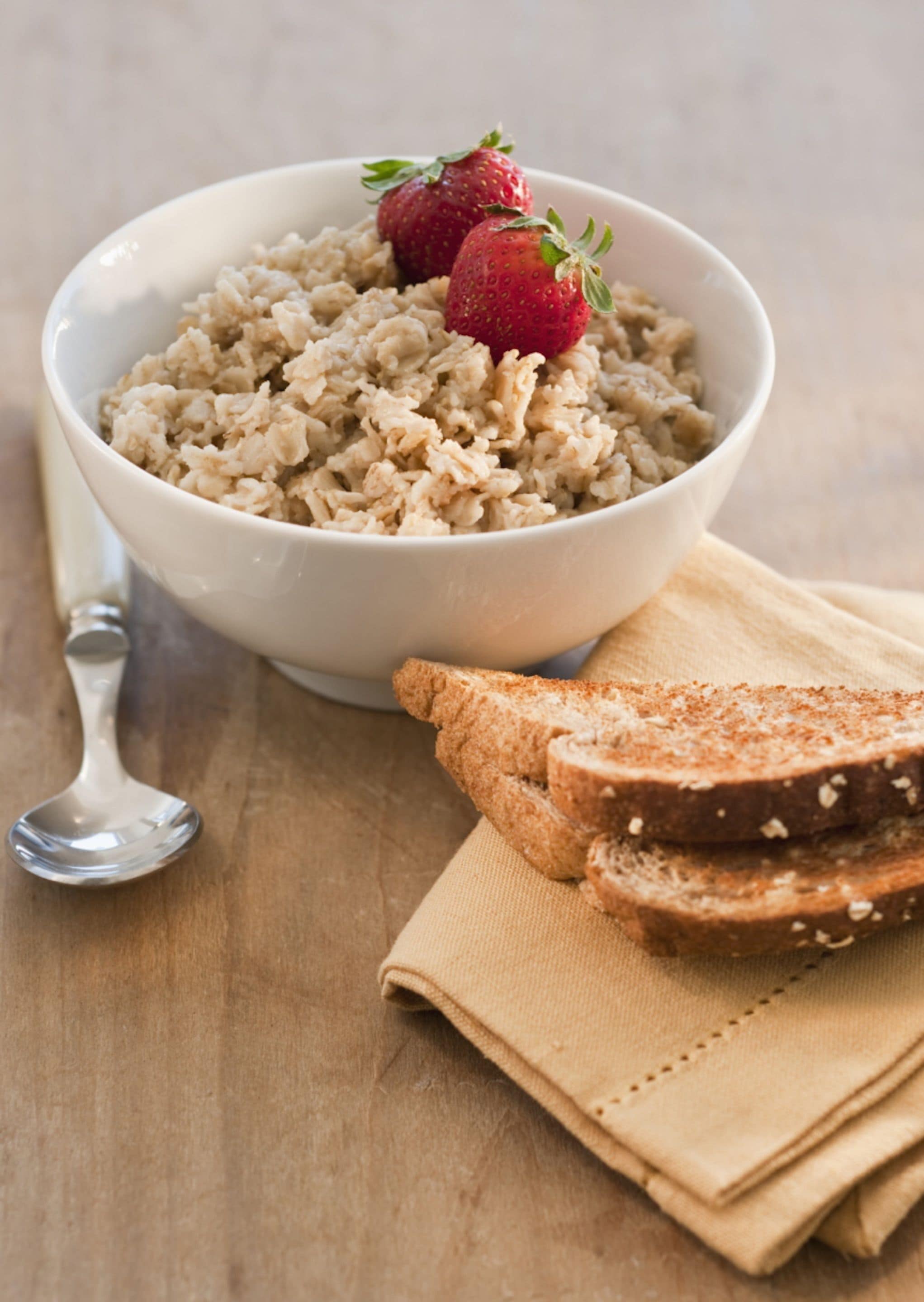 A bowl of oatmeal with strawberries on a wooden table, accompanied by a spoon and slices of bread on a napkin.