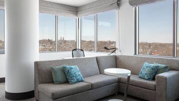Un salon moderne avec un canapé d’angle gris, deux coussins assortis, une petite table d’appoint ronde blanche et de grandes fenêtres offrant une vue sur la ville.