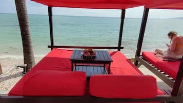 Una cabaña de playa con una mesa y sillas, una persona sentada en un banco rojo con cojín y vistas al océano.