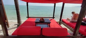 Una cabaña de playa con una mesa y sillas, una persona sentada en un banco rojo con cojín y vistas al océano.