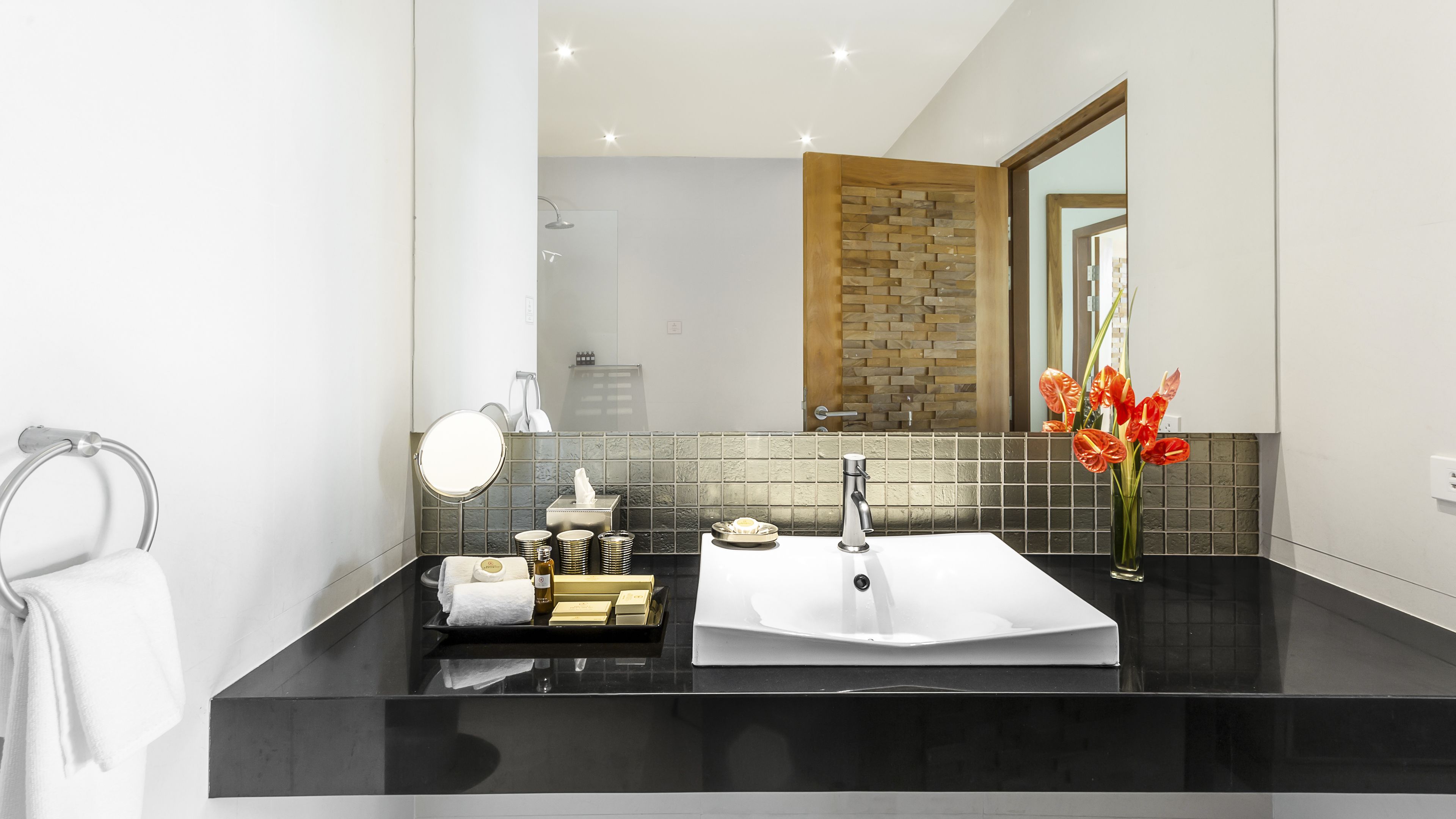 A modern bathroom with a black countertop, a white sink, a mirror, and a vase with orange flowers.