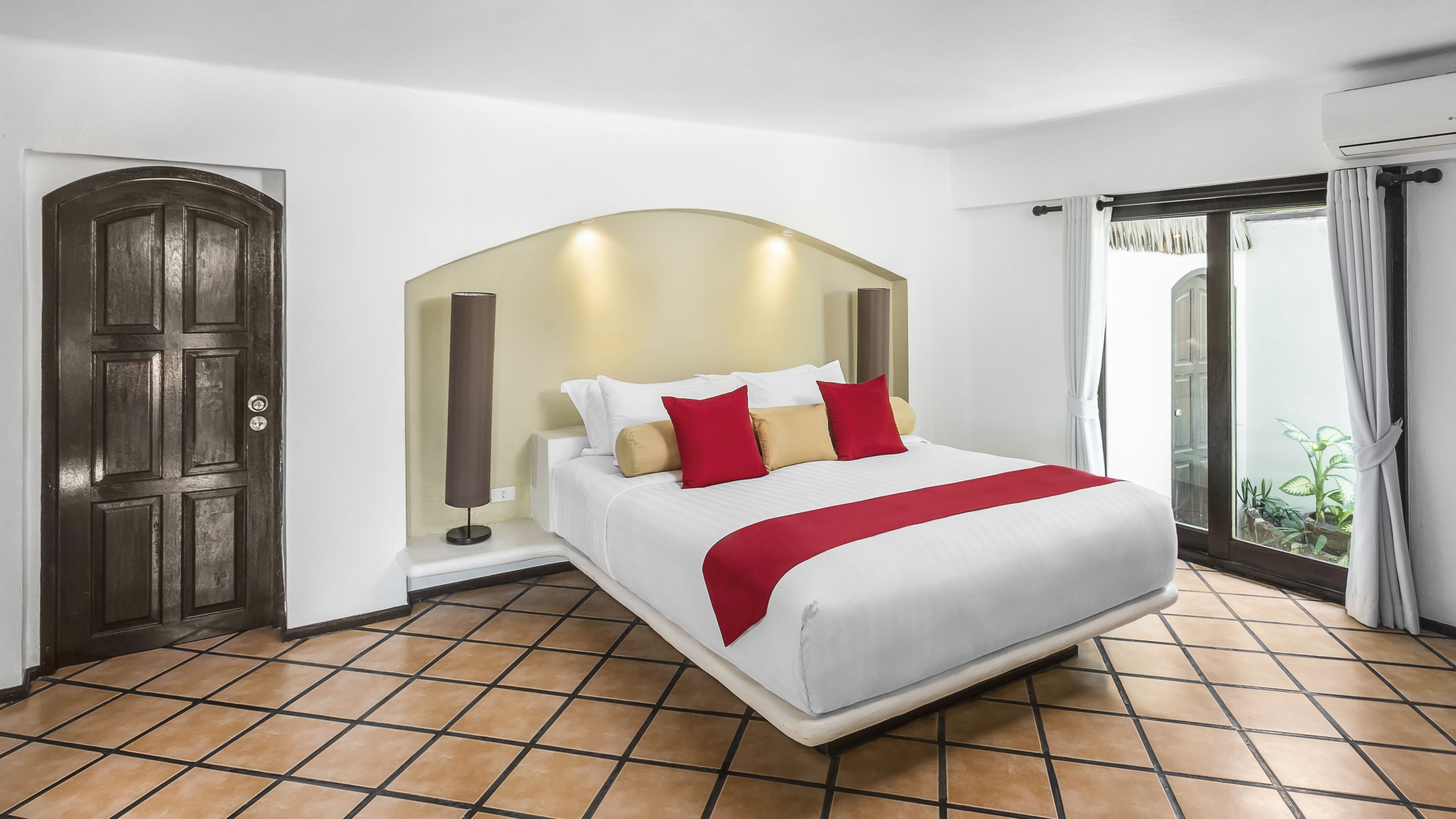 Habitación de hotel moderna con una cama grande, una puerta de madera oscura, una lámpara de pie y vistas a la vegetación por la ventana.