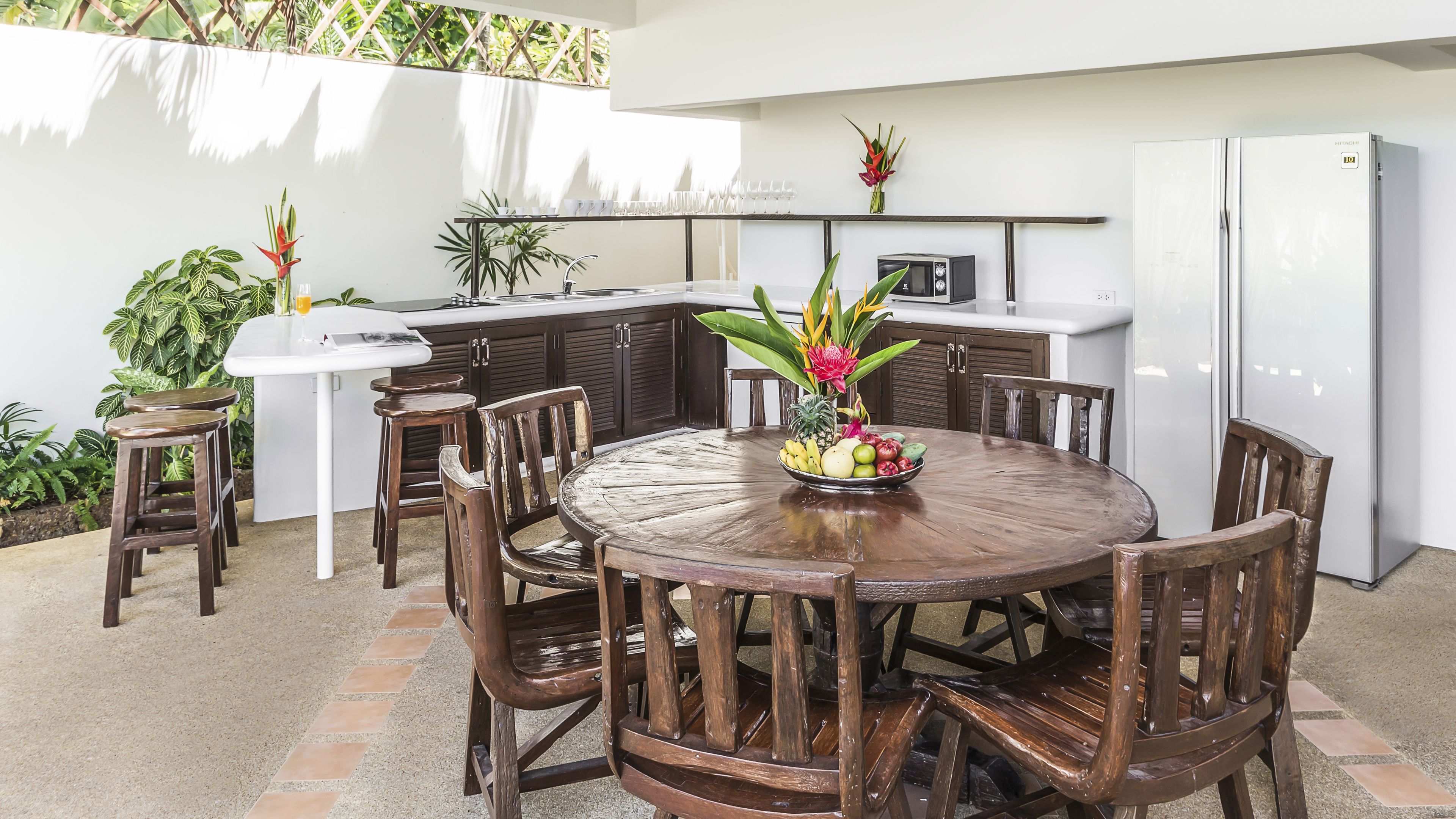 Un comedor con una mesa de madera redonda, seis sillas, un frutero y una cocina con refrigerador y microondas.