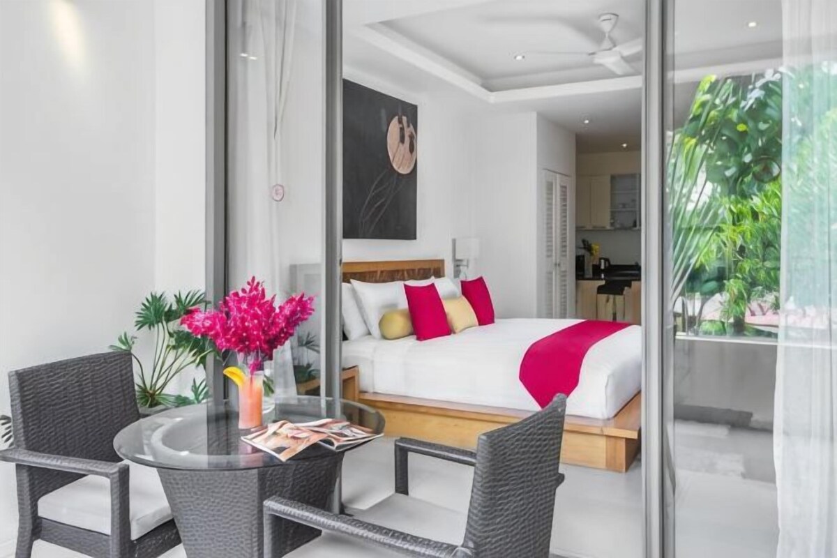 A modern hotel room with a bed, a glass table, and chairs, a view of the kitchen, and a balcony with plants.