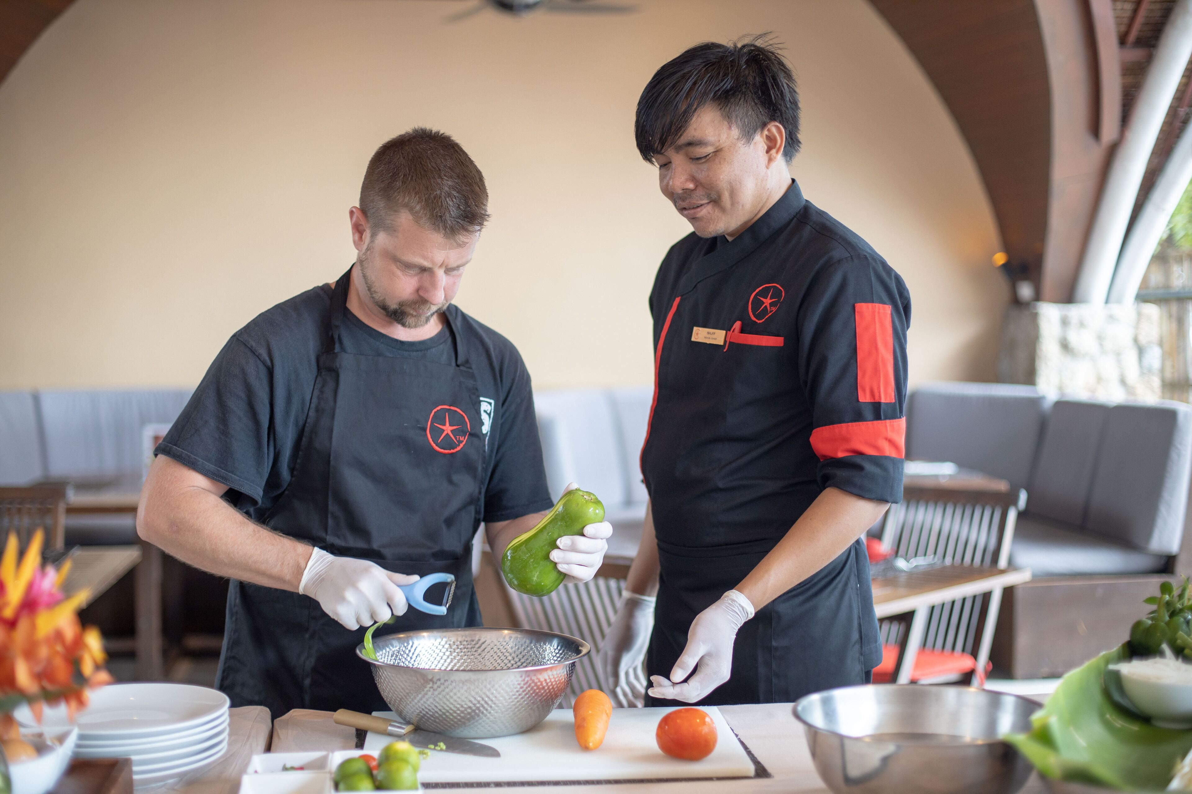 Two individuals in a kitchen preparing food, with a table full of vegetables and kitchen utensils.