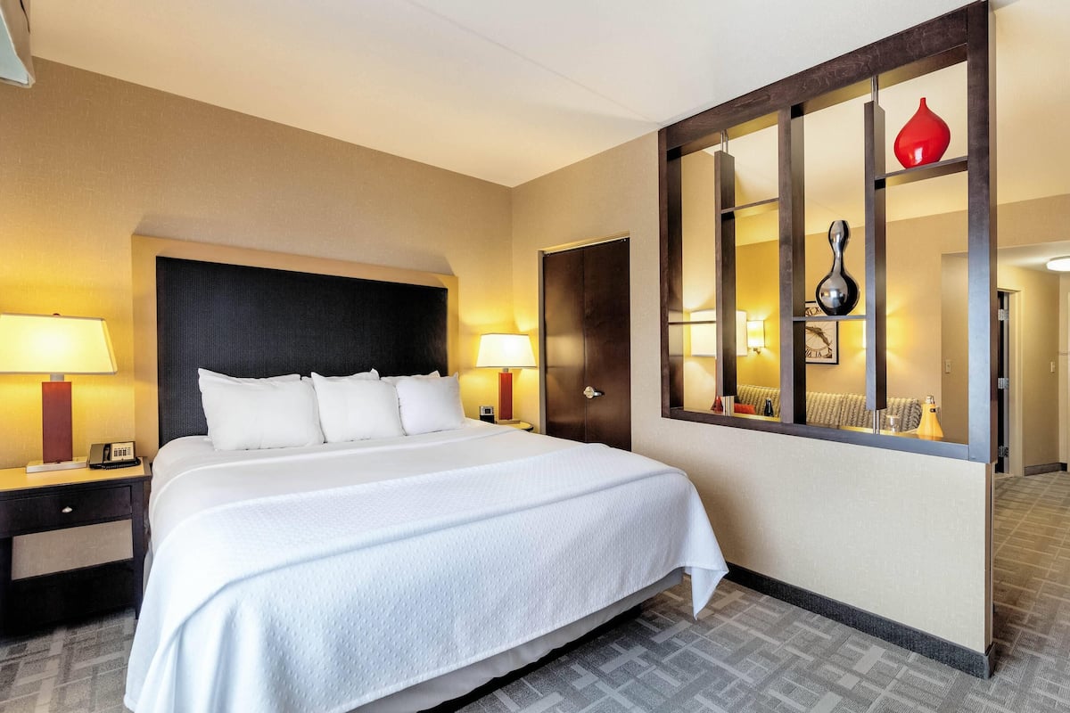 A hotel room with a large bed, bedside lamps, a wooden headboard, a mirror, and a decorative shelf with a red vase.