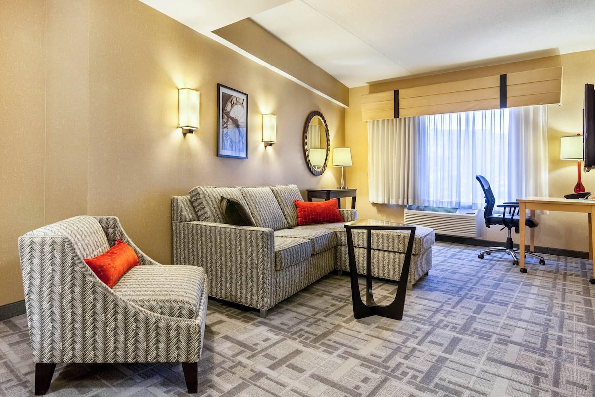 A hotel room with a sofa, armchair, and a small table. There is a window with curtains, a lamp, and a clock on the wall.