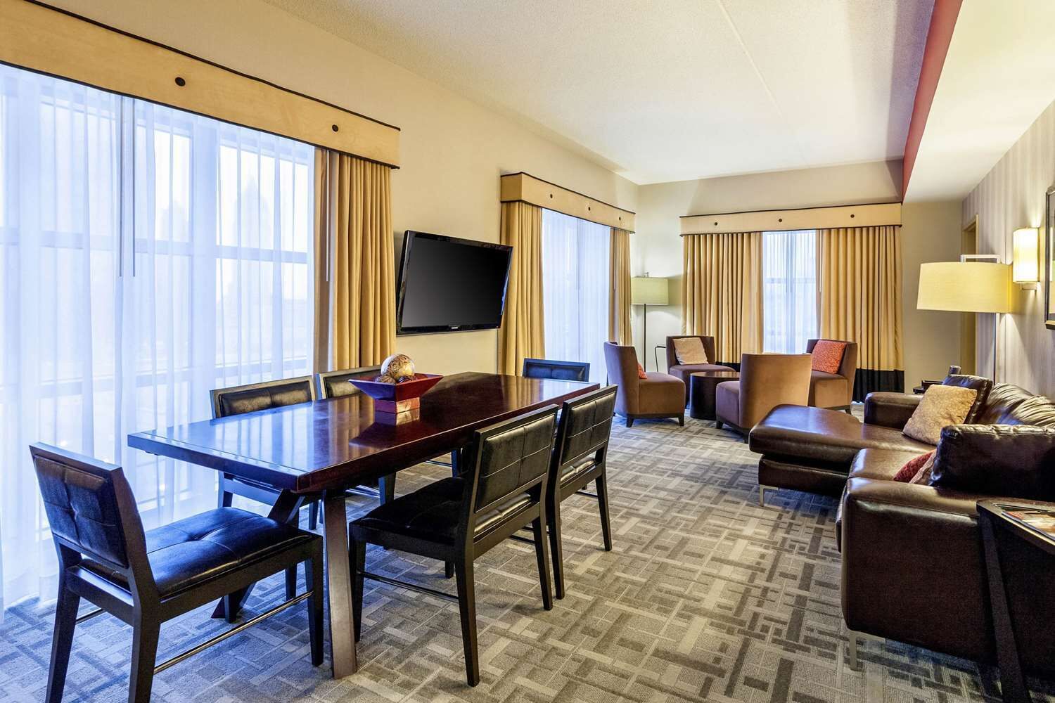 A hotel room with a dining area, a living area with a sofa and armchairs, a flat-screen TV mounted on the wall, and large windows with curtains.