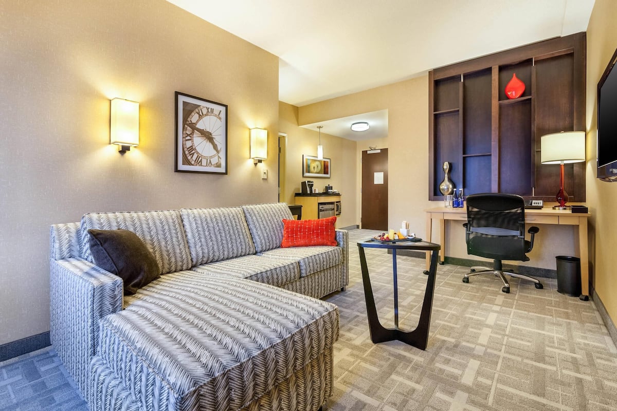 A hotel room with a large sofa, a small table, a desk with a chair, a TV, and wall-mounted lights.