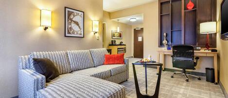 A hotel room with a large sofa, a small table, a desk with a chair, a TV, and wall-mounted lights.