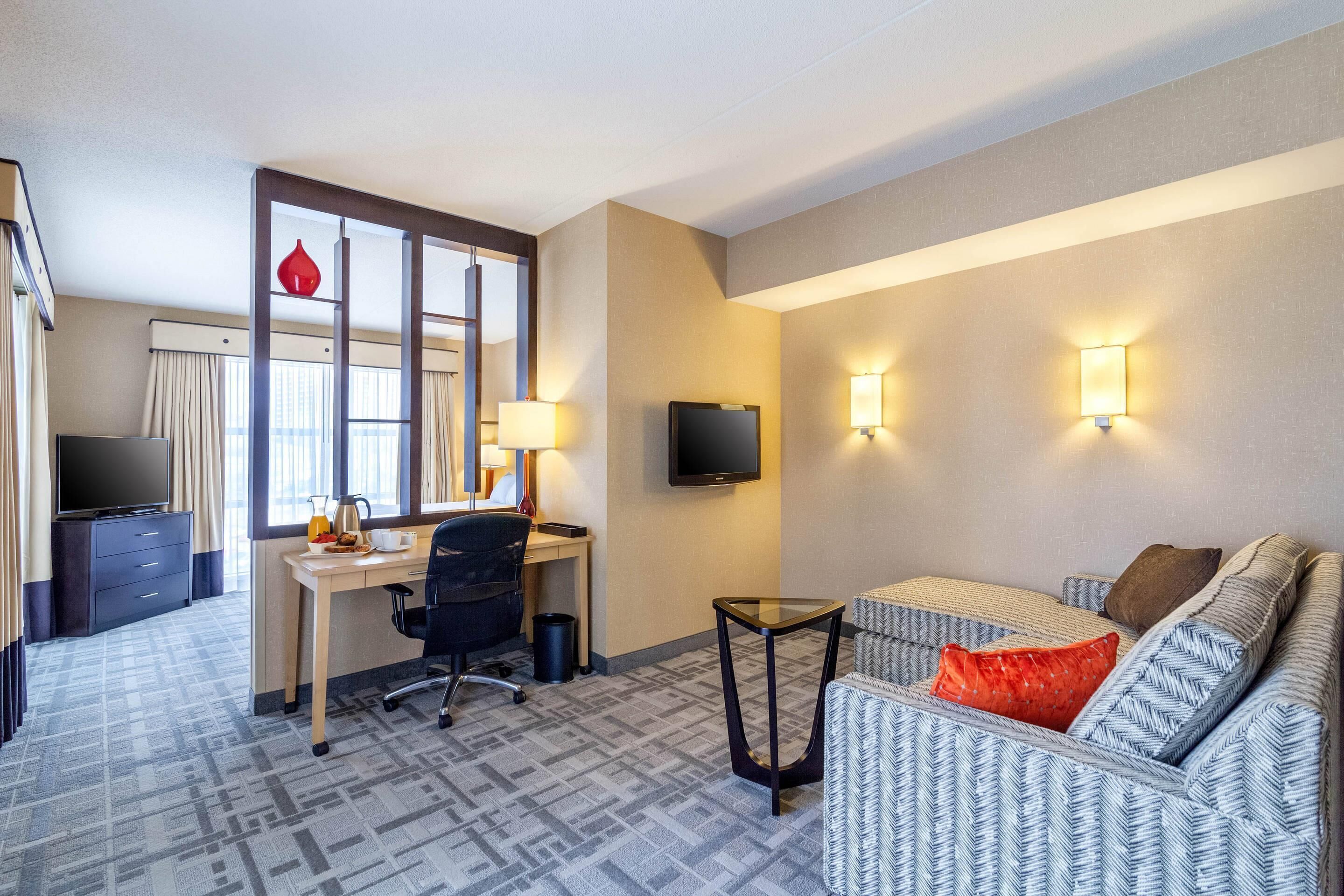 Habitación de hotel con escritorio, silla, sofá, televisor y dos luces fijadas a la pared.