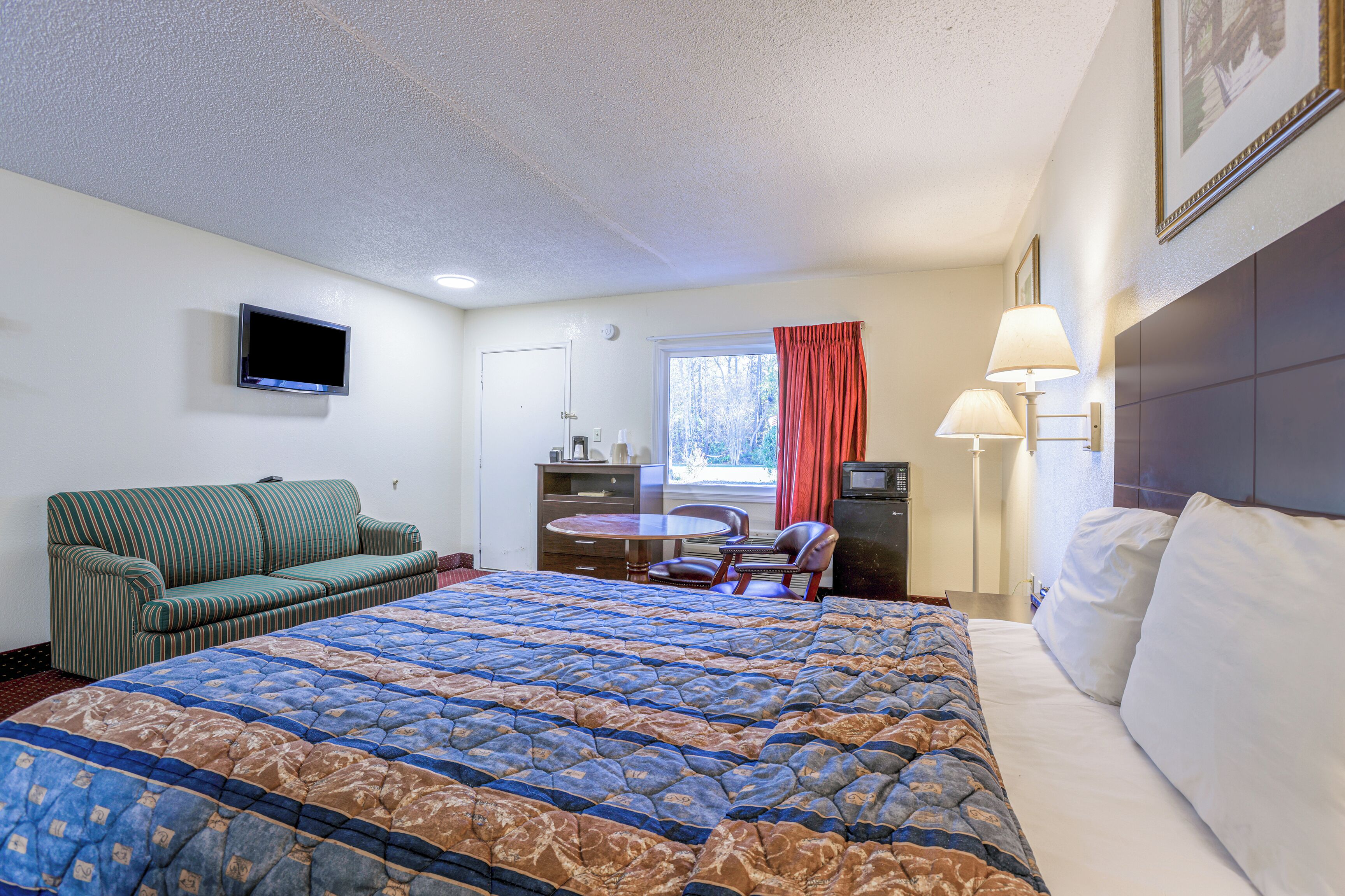 A hotel room with a large bed, a sofa, a desk with a chair, a television, and a window with curtains.