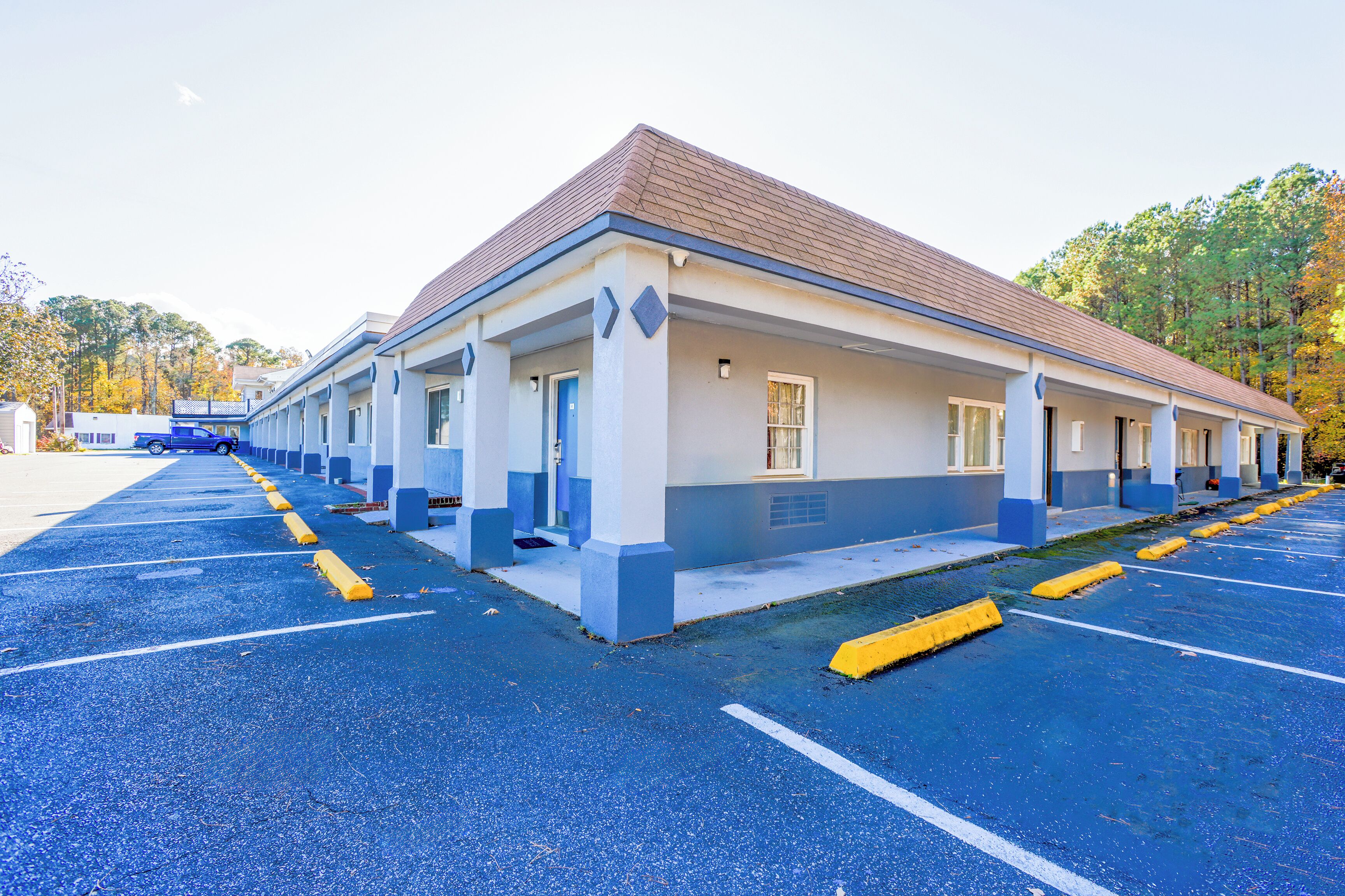 Un motel con una hilera de habitaciones, un estacionamiento techado y un amplio estacionamiento azul.