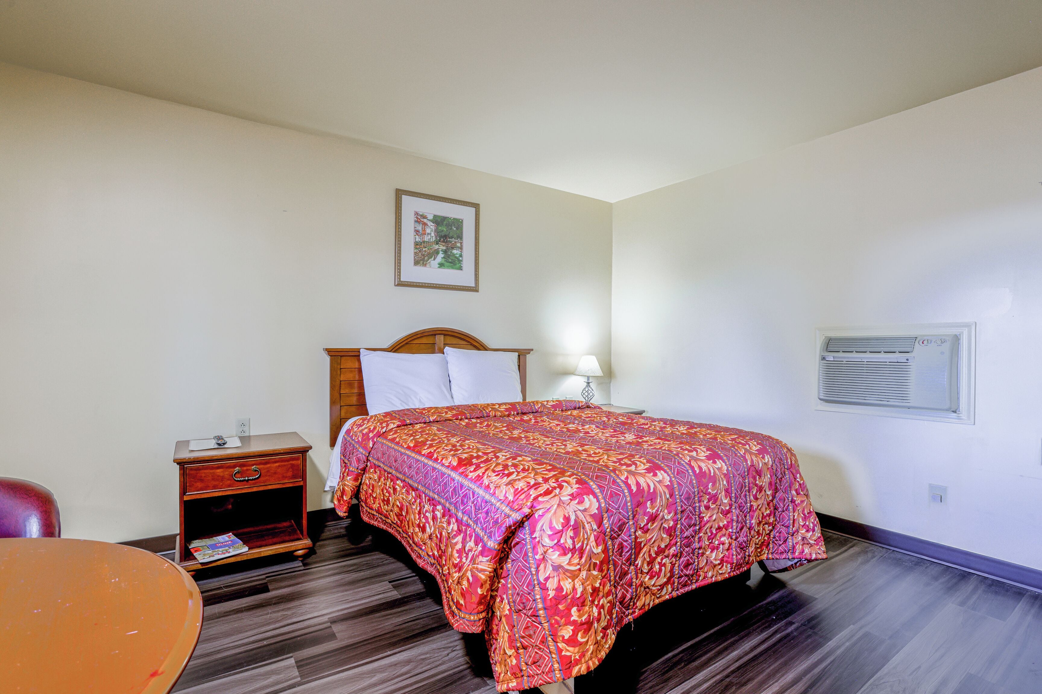 A hotel room with a bed, a nightstand, a table, and a framed picture on the wall.