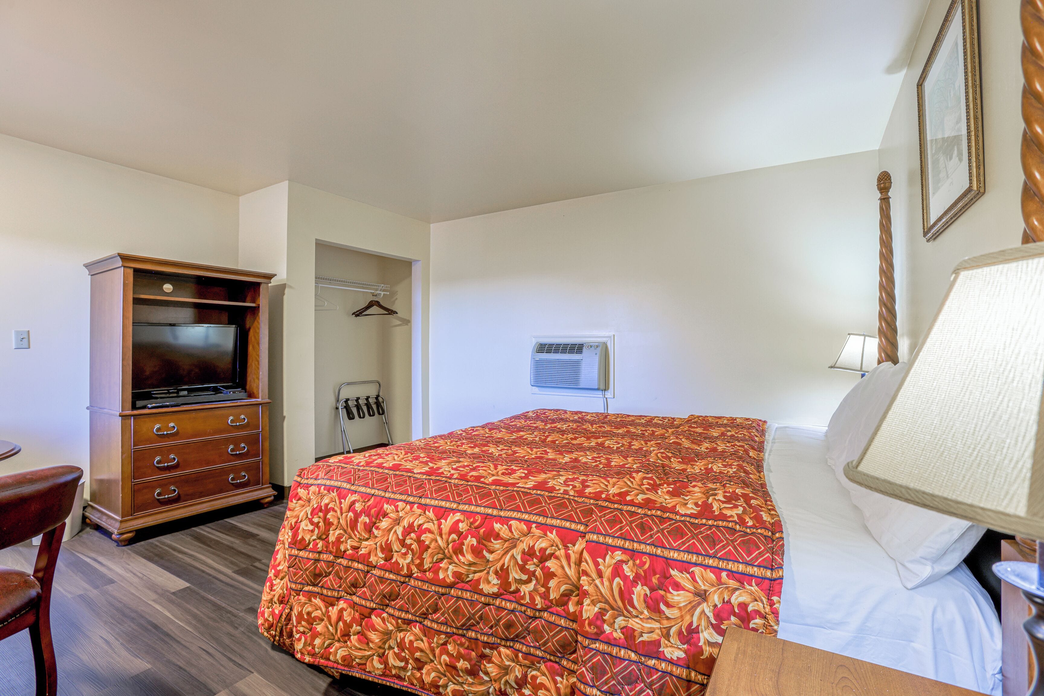 A hotel room with a large bed, a wooden dresser, a television, a wardrobe, and a bedside lamp.