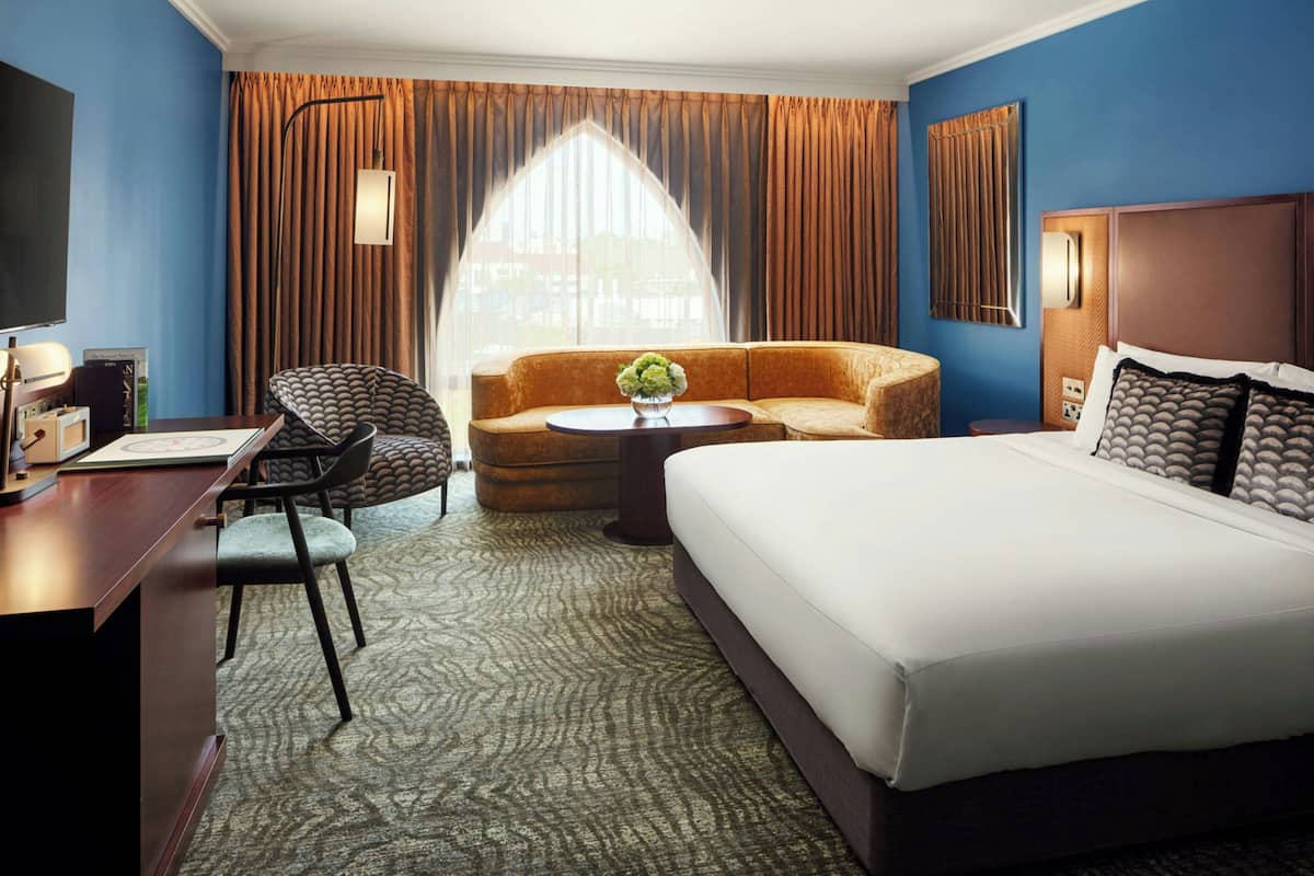 A hotel room with a large bed, a sofa, a desk with a chair, a small table with a vase, and a large window with curtains.