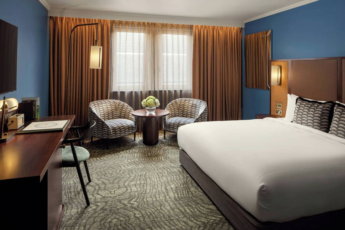 A hotel room with a large bed, two armchairs, a small table with a vase, and a desk with a lamp.