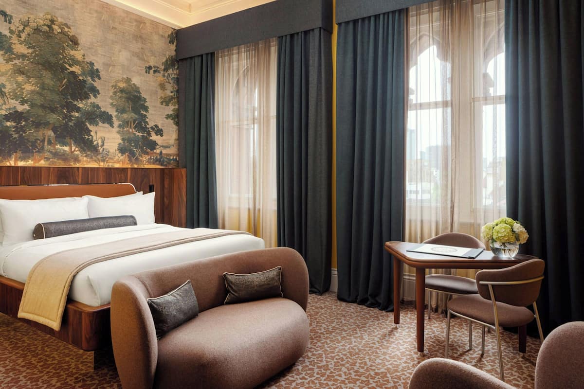 A hotel room with a large bed, a sofa, a dining table with chairs, and a view of trees through sheer curtains.