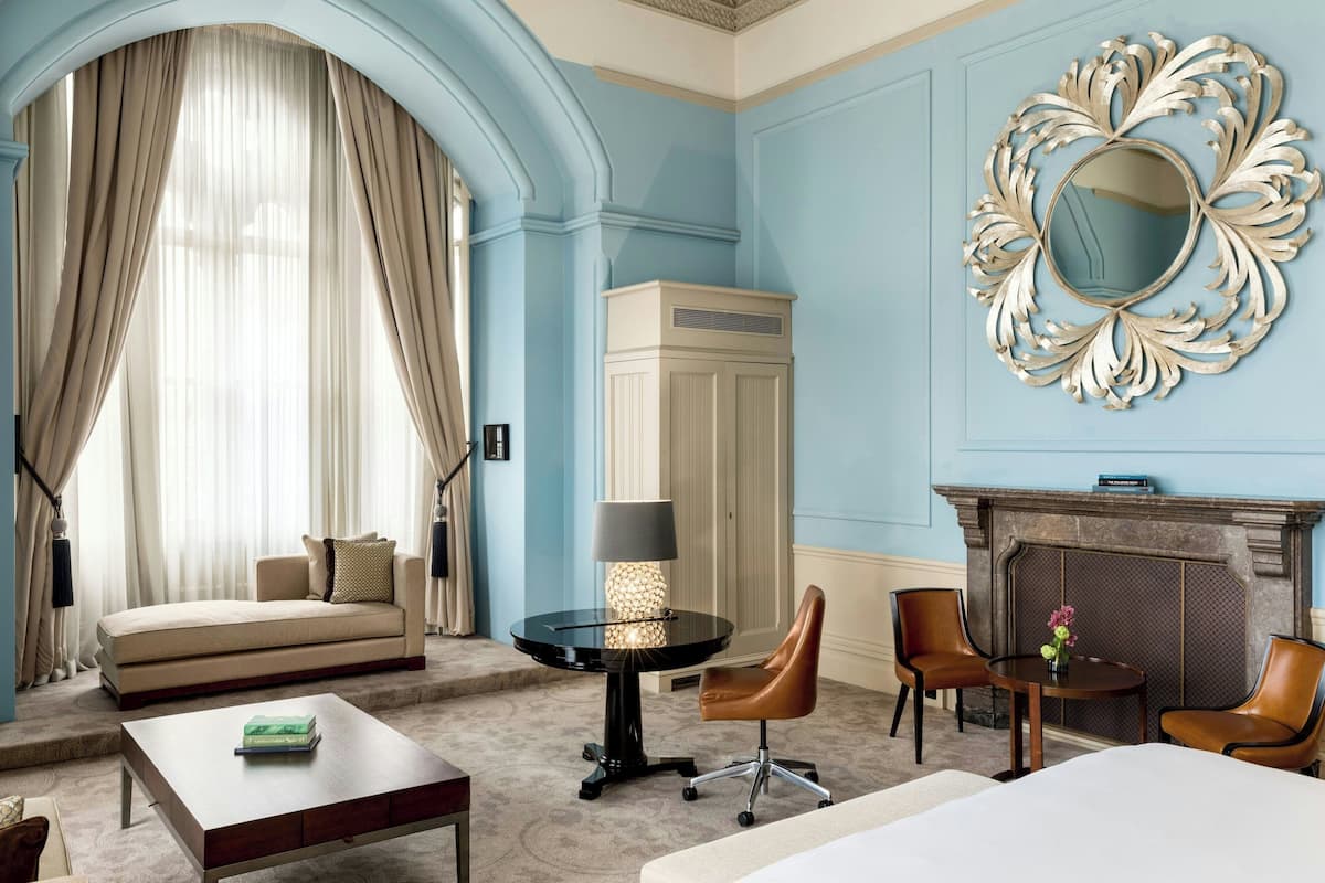 A hotel room with a sofa, a round table with a lamp, two chairs, a bed, and a fireplace.