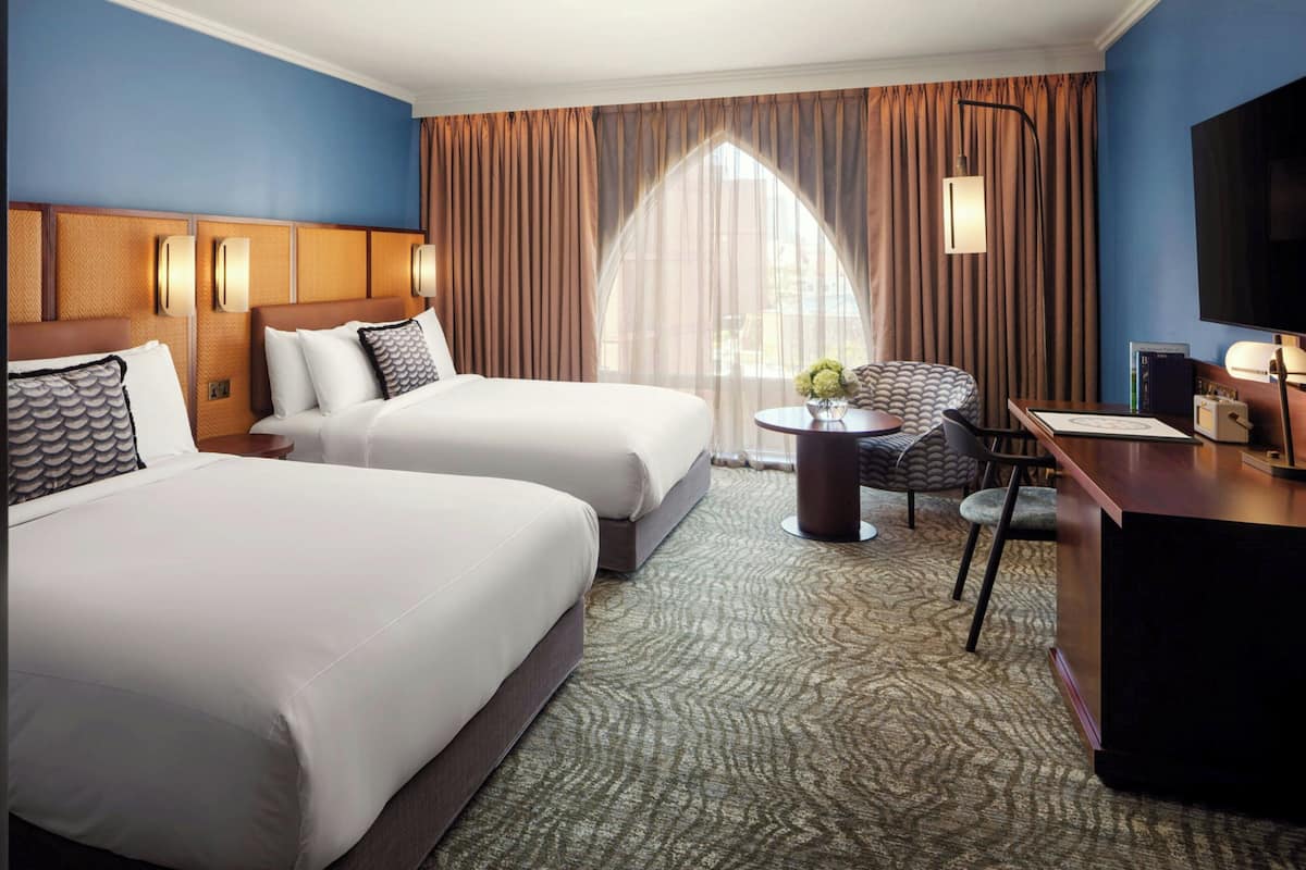 A hotel room with two beds, a desk, a chair, a television, and a window with curtains.