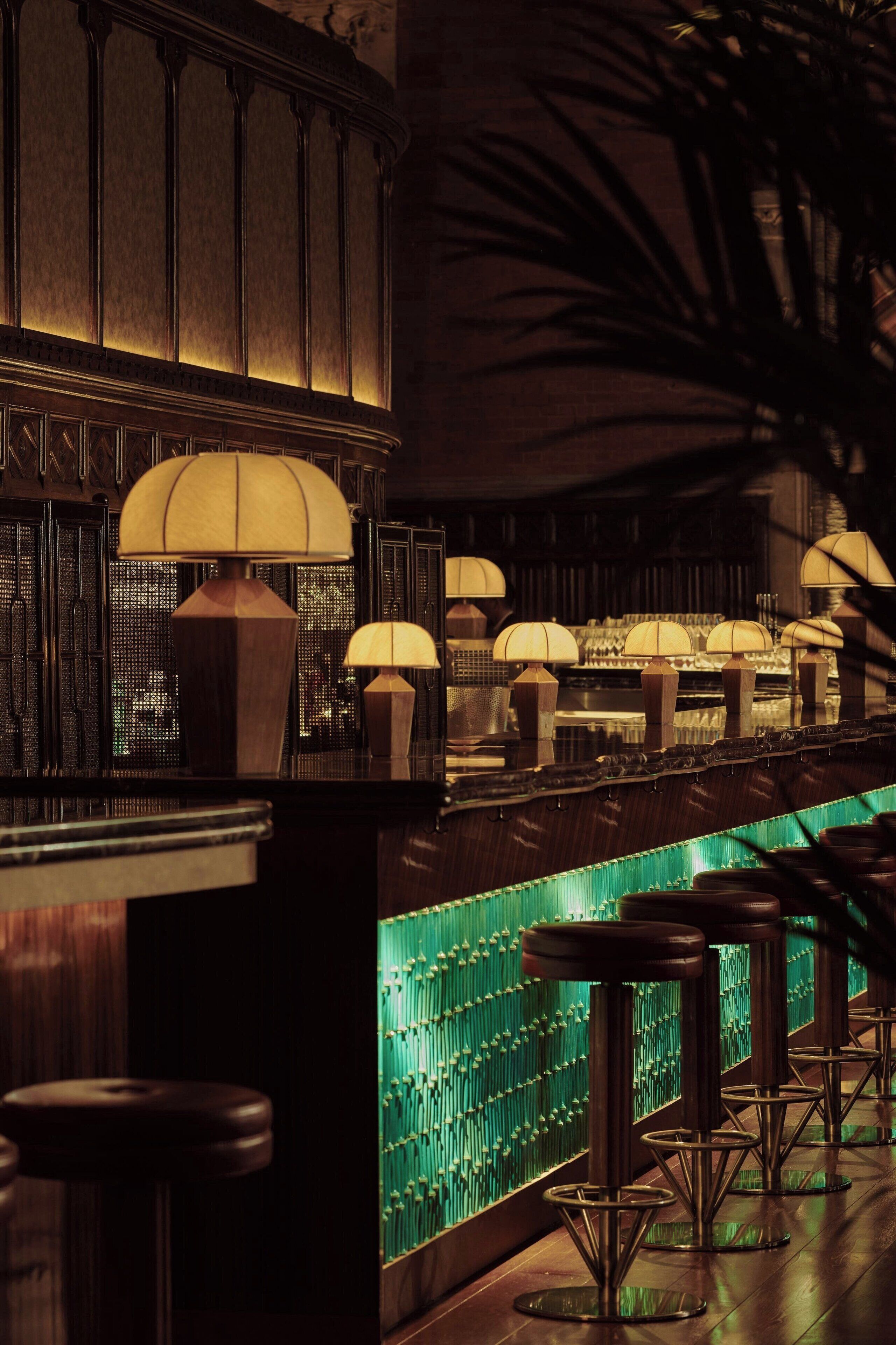 A bar area with multiple stools and lamps, a green patterned wall, and a wooden counter.