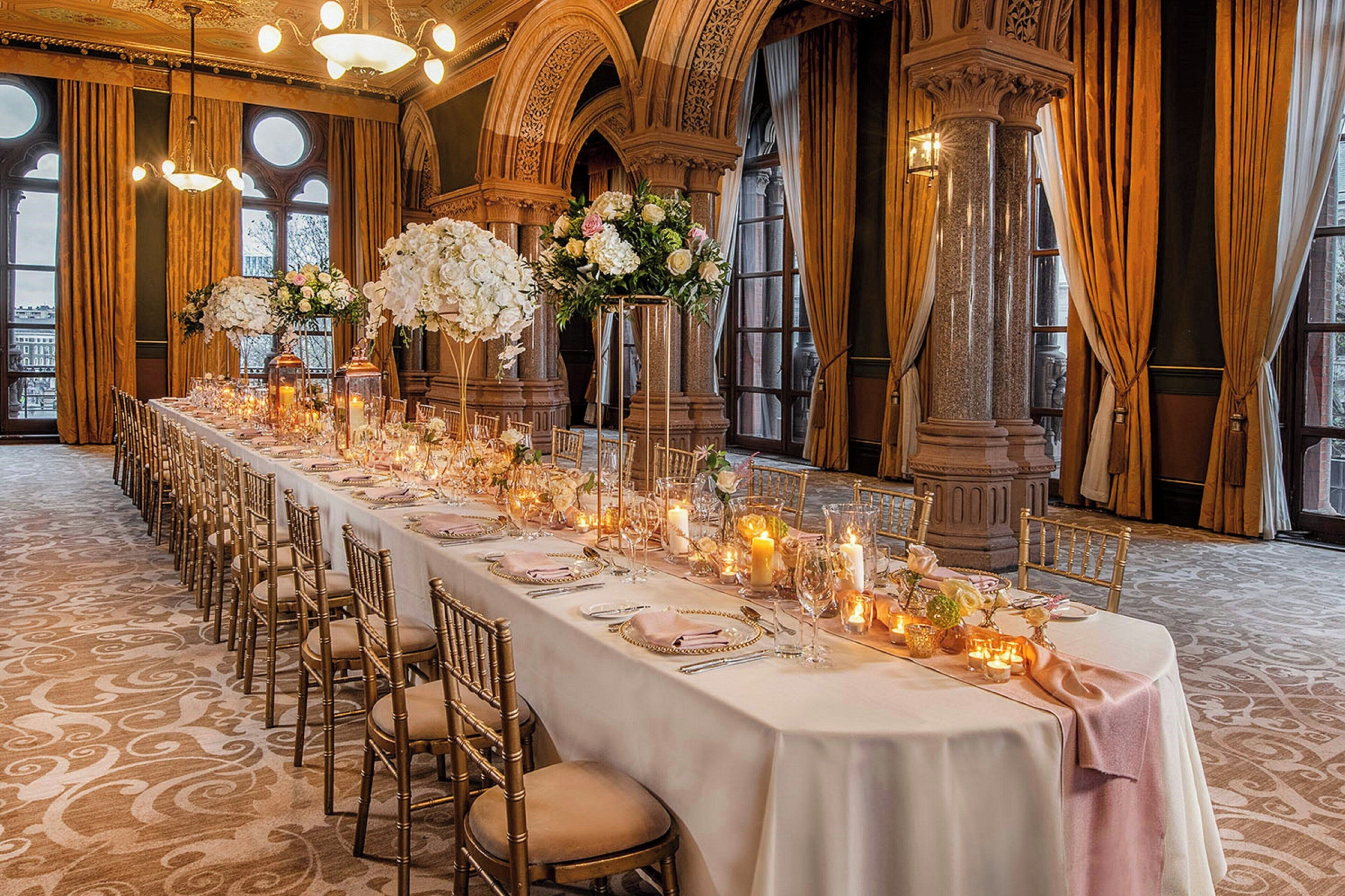 Ein langer Festtisch mit Stühlen, Kerzen und floralen Dekorationen in einem prunkvollen Saal mit Bogenfenstern und verzierten Säulen.