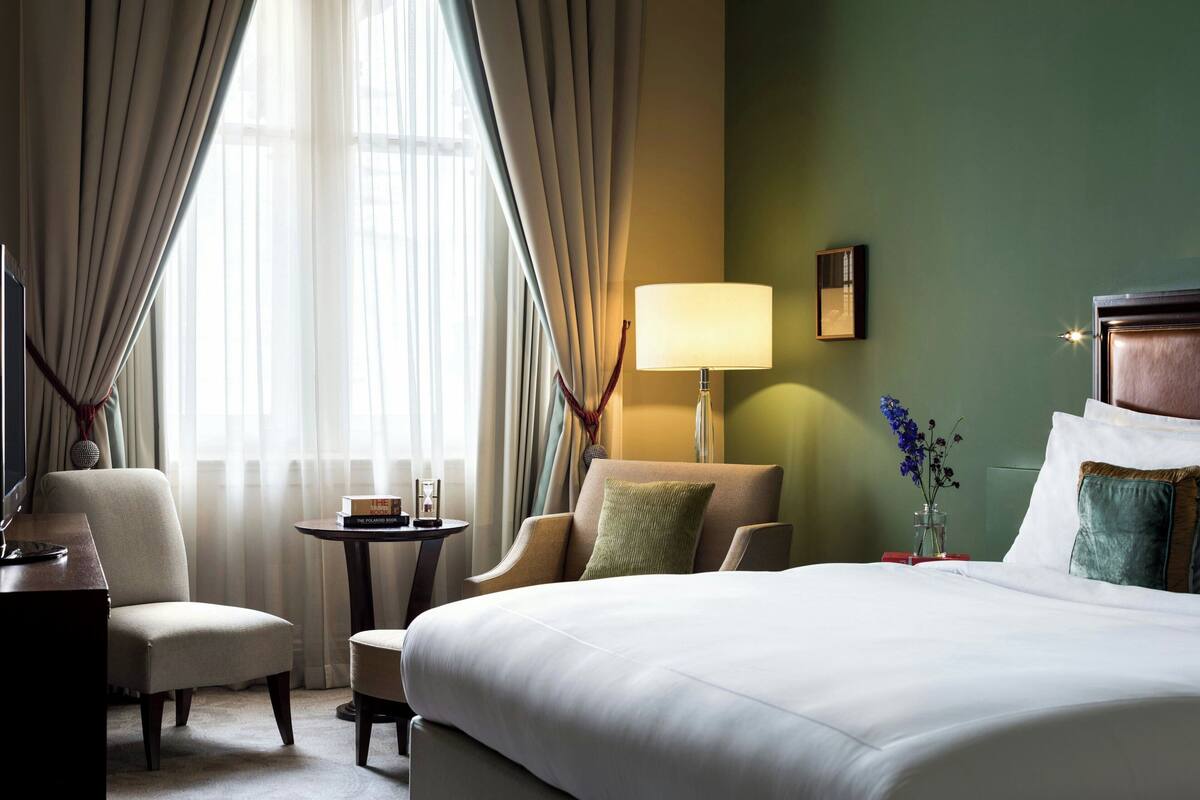 A hotel room with a large bed, a chair, a small table, a lamp, and a vase with flowers.